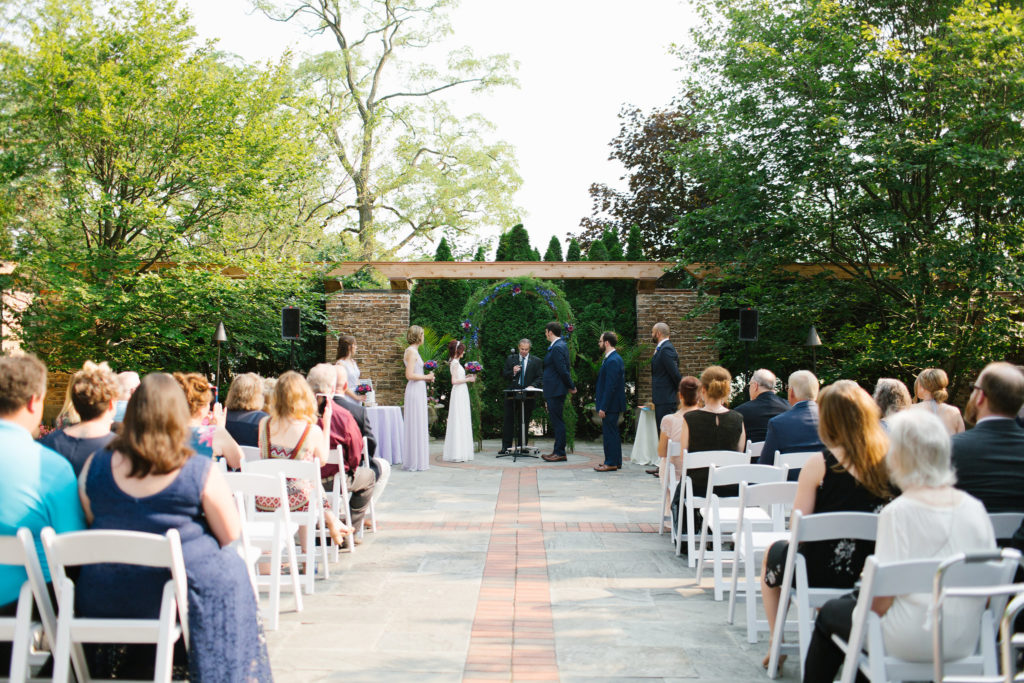 An outdoor wedding ceremony seen from behind the guests. A floral and greenery arch, brick pillars, and summer trees frame the ceremony.