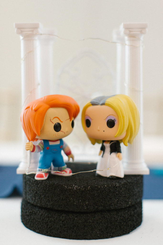 Model containing two large-headed, large-eyed humans. The one on the left has orange hair and is wearing blue overalls. The one on the right has yellow hair and is wearing a white dress. Grecian columns are in the background.