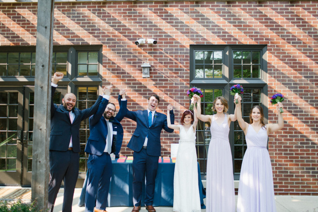 Three men in blue suits, one woman in a white dress, and two women in lavender dresses. They hold hands and hold them up in the air together with an air of celebration.