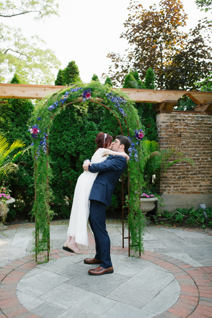 A man and a woman in front of a greenery arch. He lifts her off the ground. They are kissing.