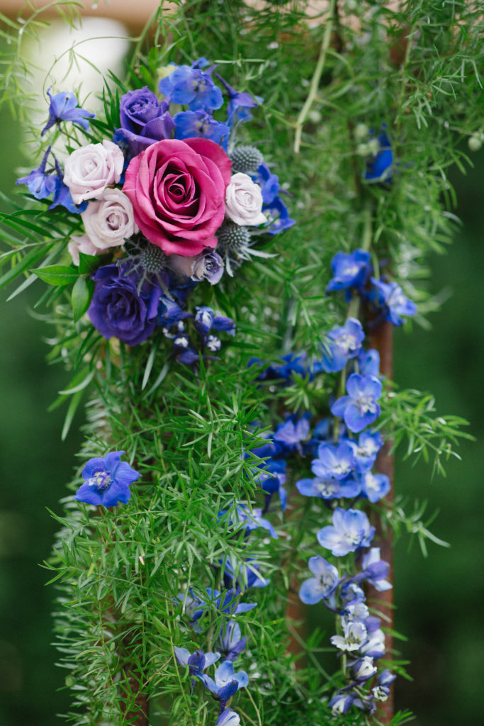 Close-up view of small-leaf greenery, dark and light pink roses, and flowers in various shades of blues and purples covering an archway.
