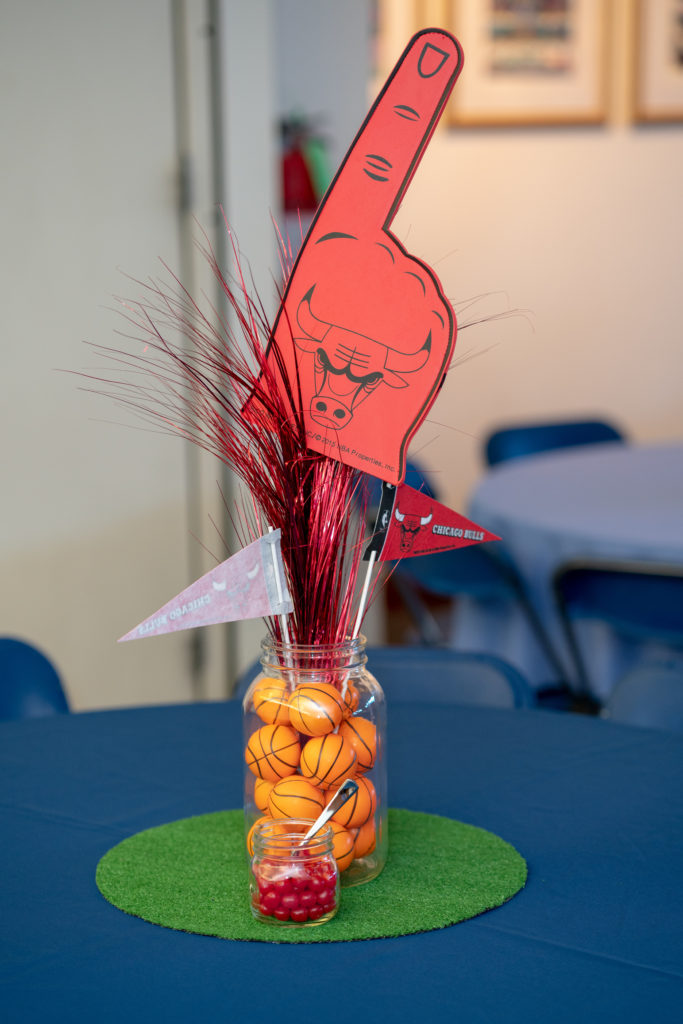 A centerpiece on a table with a blue cloth. The base is a circle of astroturf. In the middle of that is a large Mason jar filled with miniature basketballs. Two Chicago Bulls pennants and a large foam Bulls hand on a stick are standing in the jar, along with a spray of red feathery stuff. In front is a smaller jar of candy.