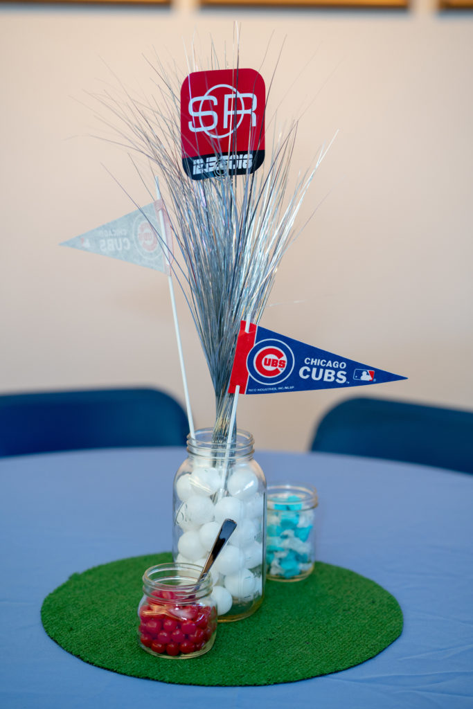 A centerpiece on a table with a blue cloth. The base is a circle of astroturf. In the middle of that is a large Mason jar filled with miniature baseballs. Two Chicago Cubs pennants are standing in the jar, along with a spray of silver feathery stuff, and a sports-looking logo saying "SR. 12-15-18." On either side are smaller jars of candy.