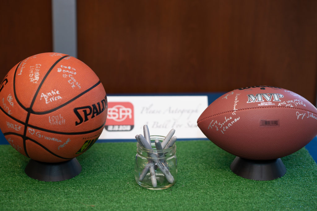 A basketball and a football on stands on a square of astroturf, along with a jar of silver Sharpie pens. People have written their names on the balls.