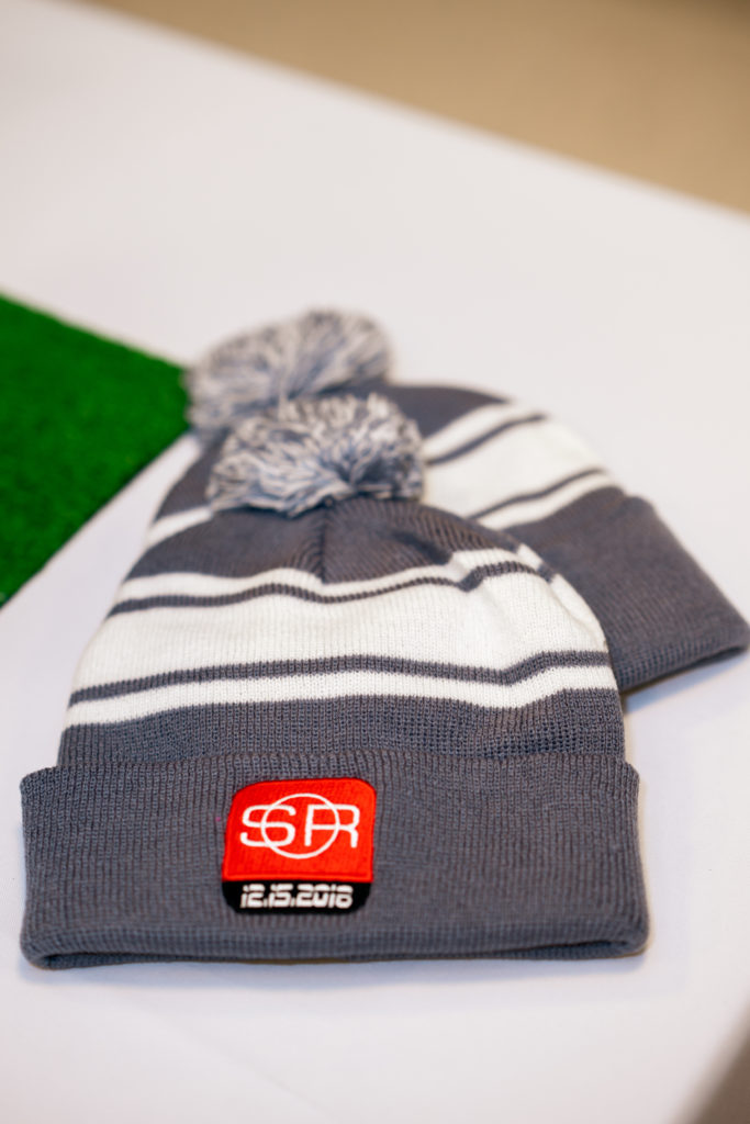 Two gray and white striped winter hats on a table. The hats have a red and white sports-themed logo saying "SR. 12-15-18."