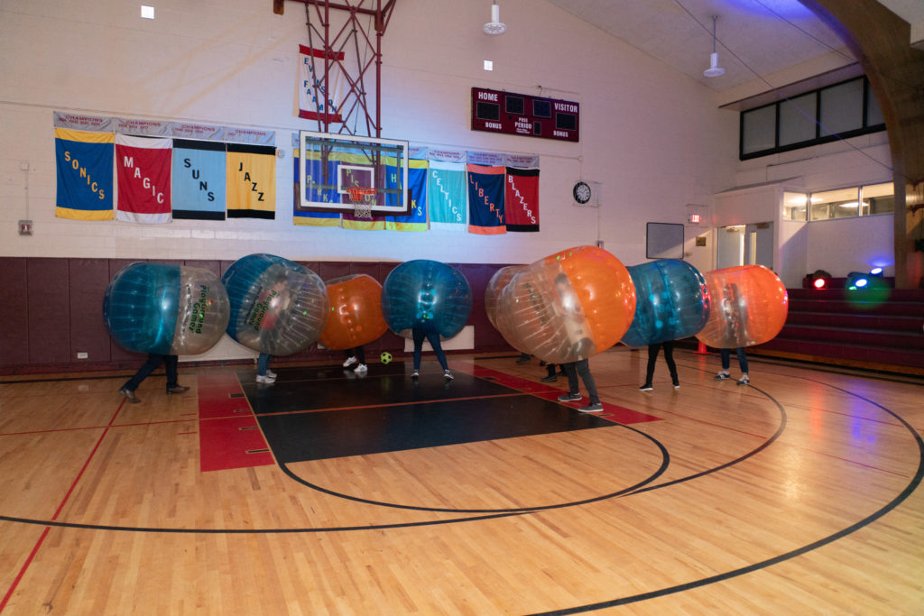 People wearing large, inflated plastic balls and playing soccer in a gym.