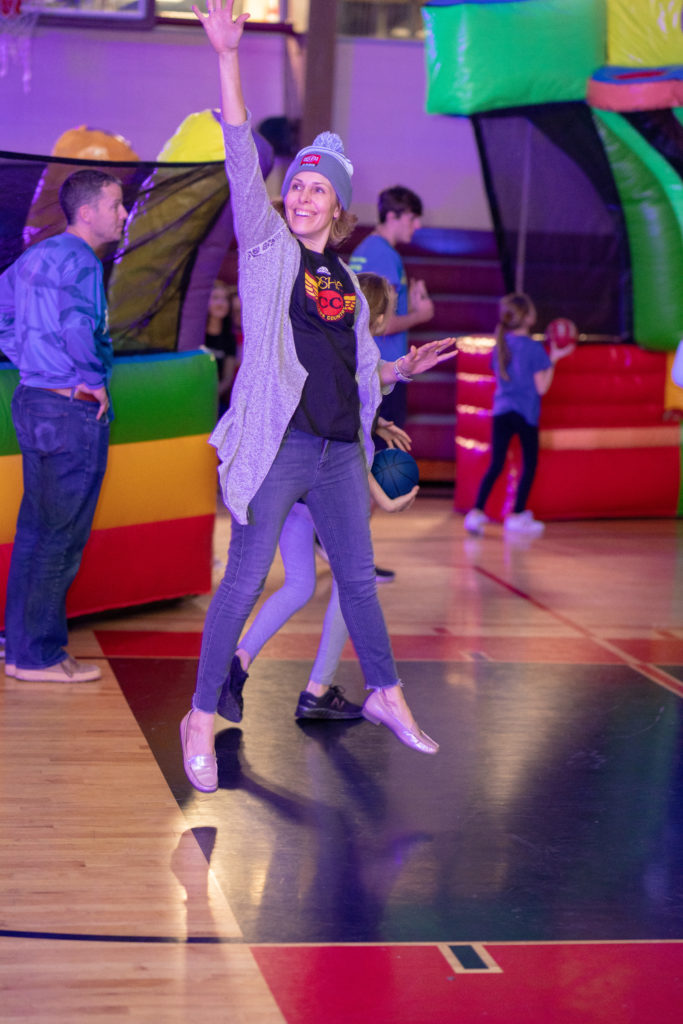 An adult in a gym full of games, jumping in the air with her arm over her head and smiling.