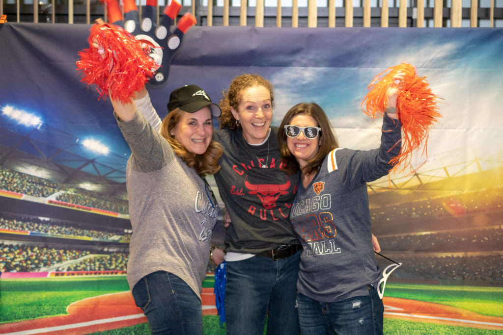 Three people with orange pom-poms and other sports gear pose in front of a backdrop showing Wrigley Field.