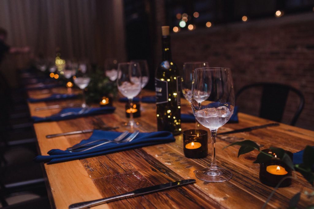 A long wooden table set with blue napkins, clear water glasses, and tea lights in cut-off colored glass bottles.