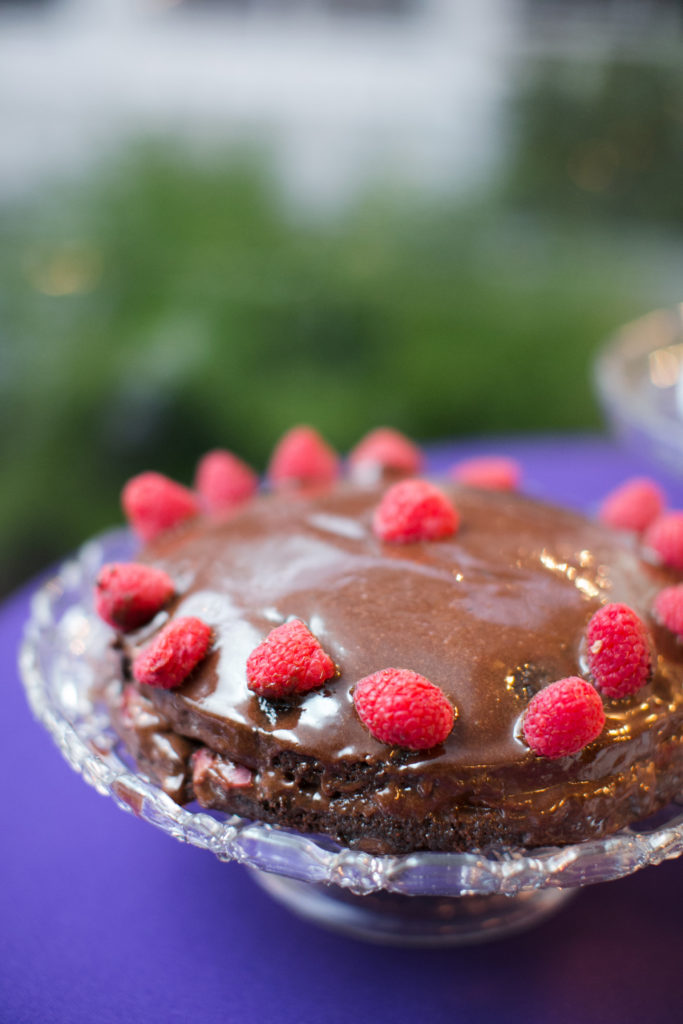 A round, single-layer chocolate cake decorated with raspberries on a clear cake stand.