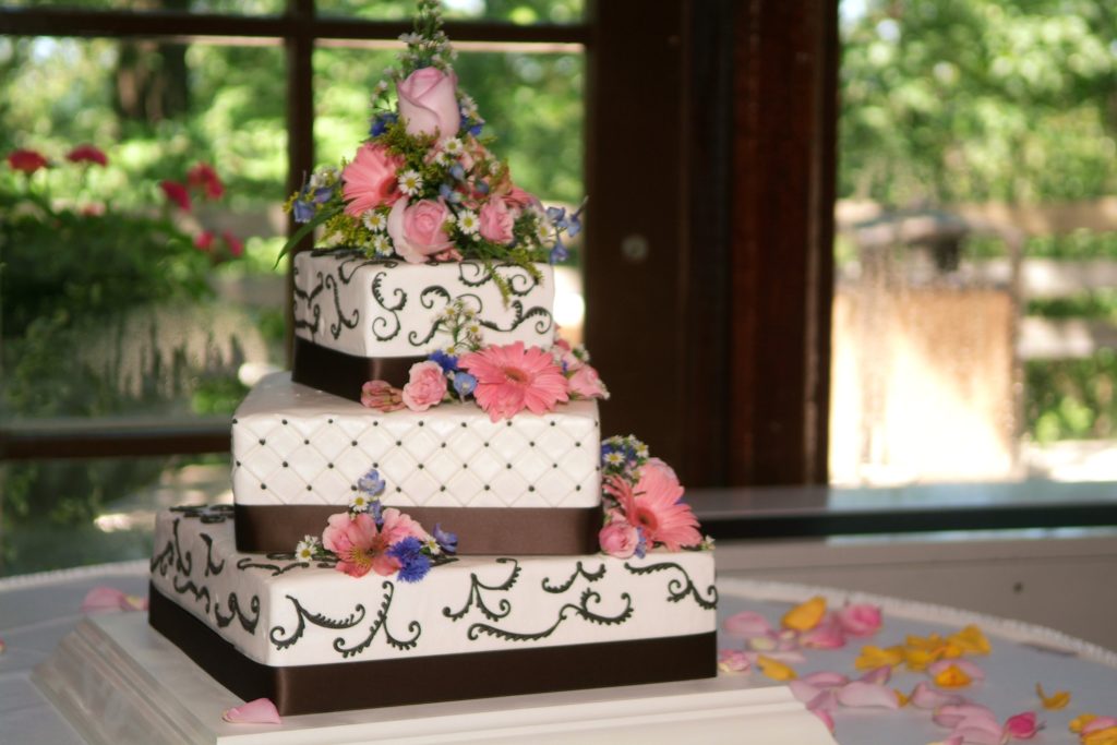A 3-tier wedding cake. The tiers are square and each one is offset 45 degrees from the one below. It is frosted in white and brown with intricate decorations, and adorned with pink flowers.