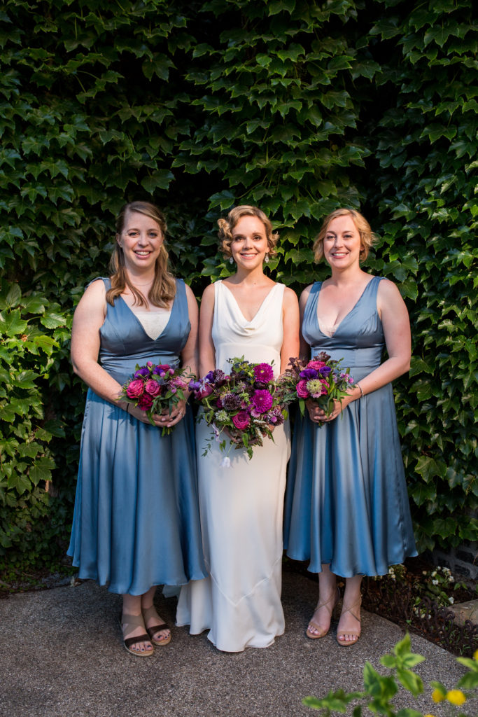 A woman in a white dress flanked by two women in blue dresses. All three hold large bouquets of flowers in reds and purples.