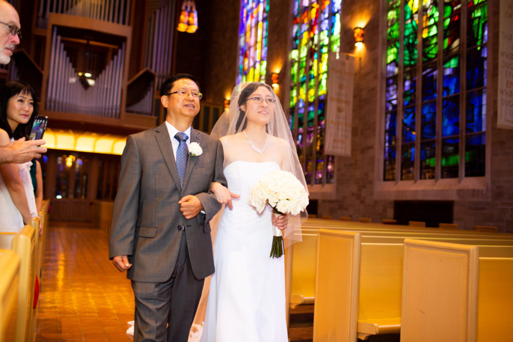 A woman wearing a white dress and a veil, and carrying a bouquet of white roses, walks down the aisle on the arm of a man in a gray suit.