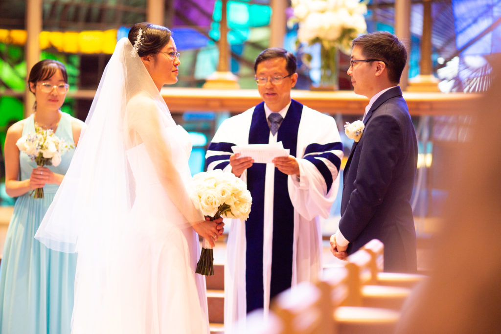 A woman in a white dress and veil, holding a bouquet of white roses, faces a man in a blue suit. Between them is a man in a clerical robe.