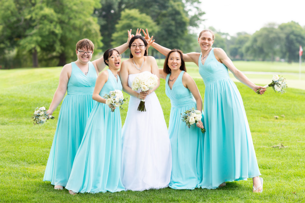 One woman in white is flanked by four women in aqua dresses. The four make "rabbit ears" over the one woman's head.