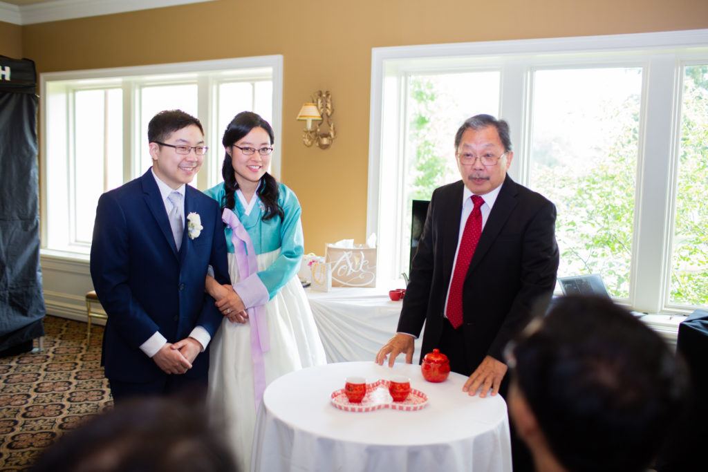 A woman in a traditional Korean dress and a man in a blue suit stand near an older man with his hands on a small table containing a red and white Chinese tea set.