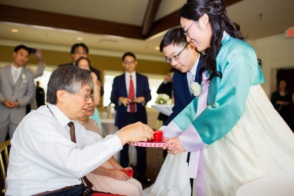 A woman in a traditional Korean dress and a man in a blue suit serve tea to an older couple.