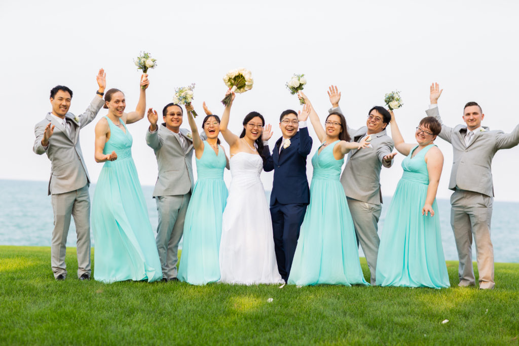 A woman in white and a man in a blue suit are flanked by people in aqua dresses and gray suits. They all have their hands in the air in a celebratory manner.