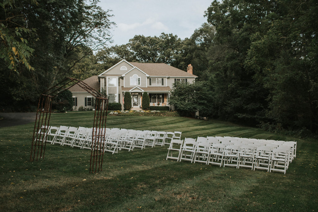 A large lawn in front of a suburban house, with rows of white chairs set up on two sides of an aisle. A rustic-looking arch is in front of the chairs.