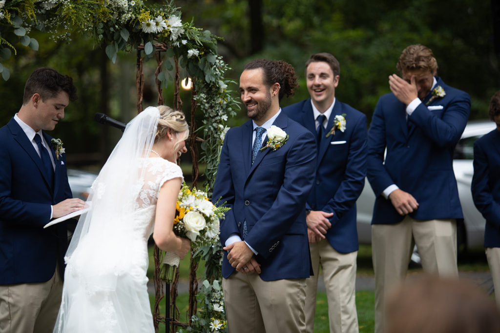 A bride and groom in front of a rustic, flower-festooned arch, with an officiant and groomsmen in the background. The bride is leaning forward laughing, while the groom and a groomsman smile.