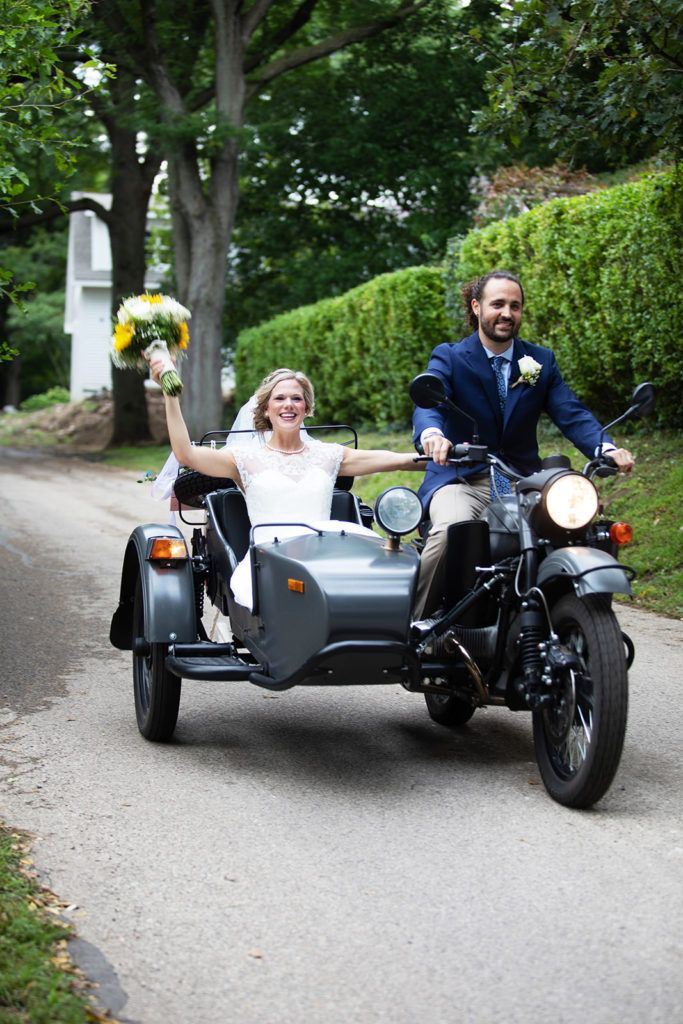 A man in a blue jacket and khaki pants, with a white boutonniere, rides a motorcycle with a sidecar. In the sidecar is a woman in a white dress and veil, holding up a bouquet of yellow and white flowers. Both are smiling.