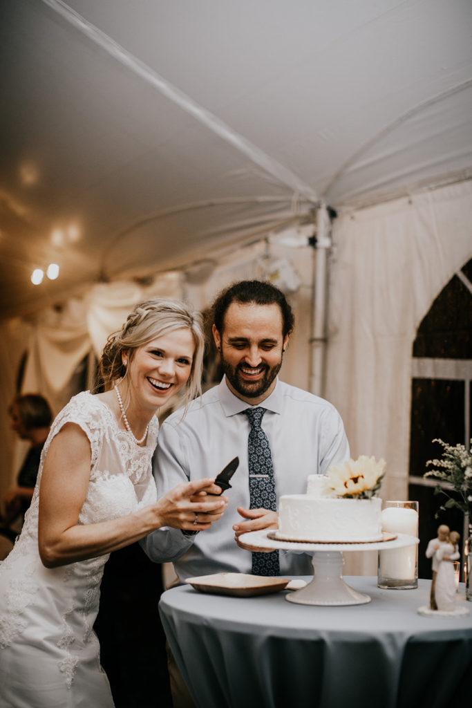 Bride and groom in a large reception tent cutting a cake with a hunting knife and laughing.