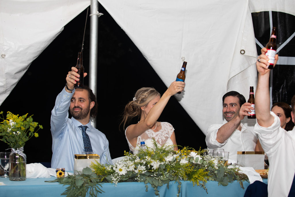 Several people sit at a long table covered in a blue cloth with vases and garlands of yellow and white flowers on it. All are raising beer bottles for a toast.