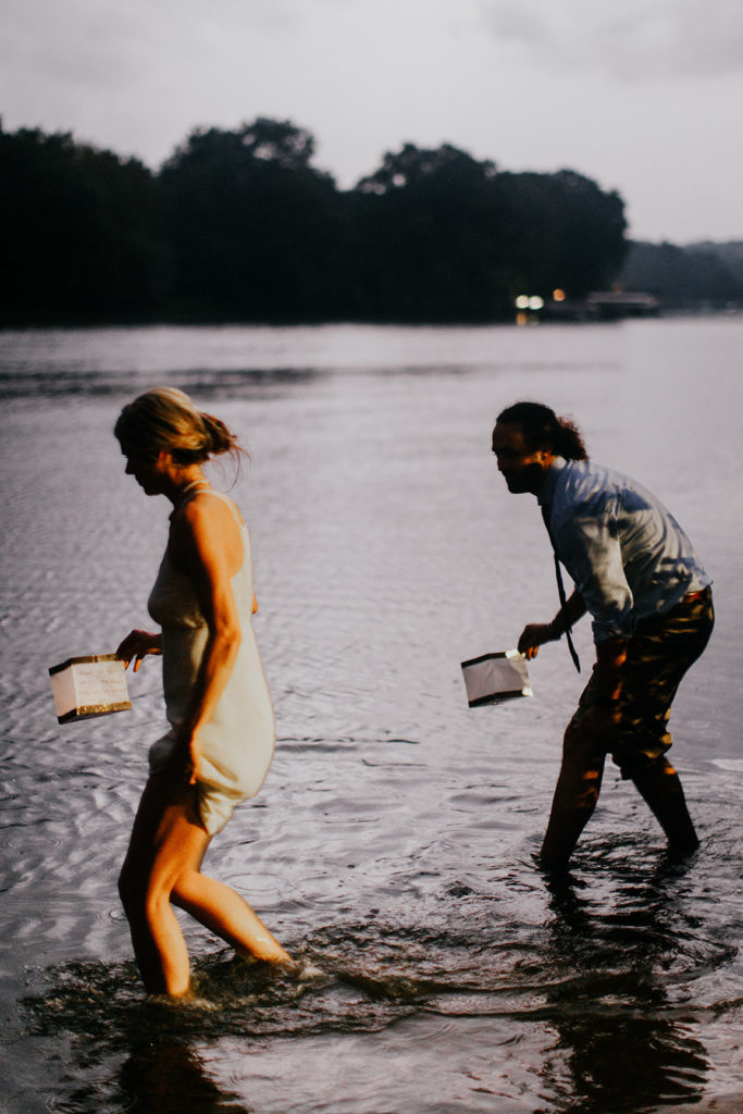 A man and a woman, with their clothes hiked above their knees, stand in a river. Each has a floating lantern in their hands. 