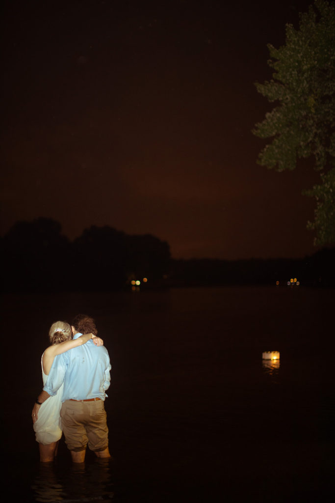 A man and a woman, seen from behind, embracing while they stand in a river above their knees. They are watching two lighted floating lanterns as they float downstream in the dark. 