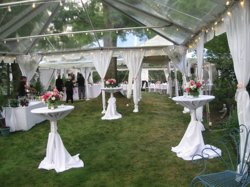 The interior of a large, clear-top tent with white leg drapes and cafe lights along the ceiling. In the foreground are three high-boy tables with white tablecloths, each one holding a large centerpiece of pink flowers.
