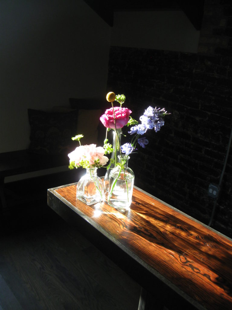 Three clear glass bud vases of various heights holding flowers in shades of pink, blue, and yellow. They are on a narrow, dark wood table and glowing in the sunshine, against a dark background.
