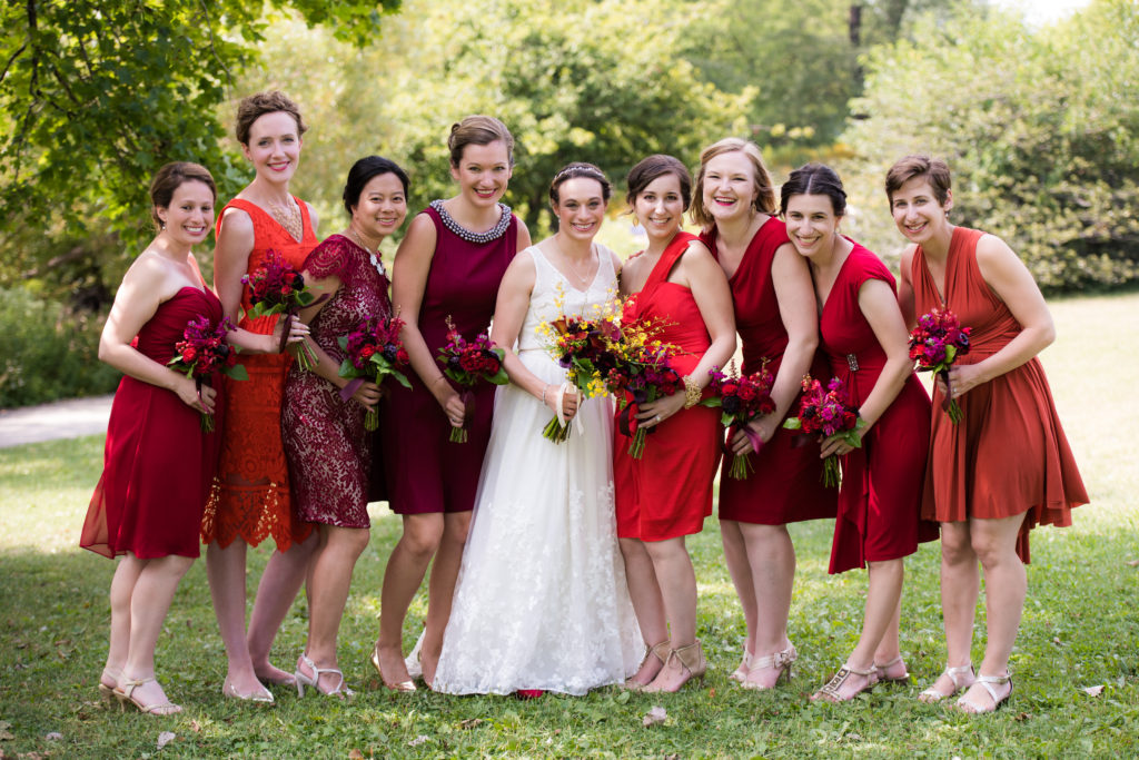 A woman in a long white dress is flaked by eight women in short dresses in various shades of red and maroon. They all hold bouquets in shades of red and maroon.