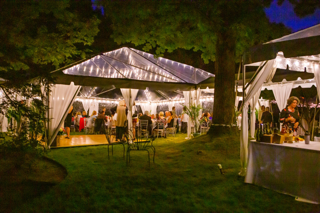 A lighted, clear-top tent on a lawn with party guests at the tables in it.