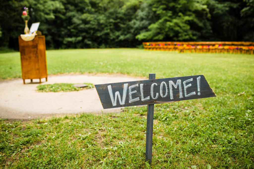 In the foreground, a rustic sign saying, "Welcome." In the background and out of focus, a large lawn bordered by forest with chairs set up for a wedding against the forest wall.