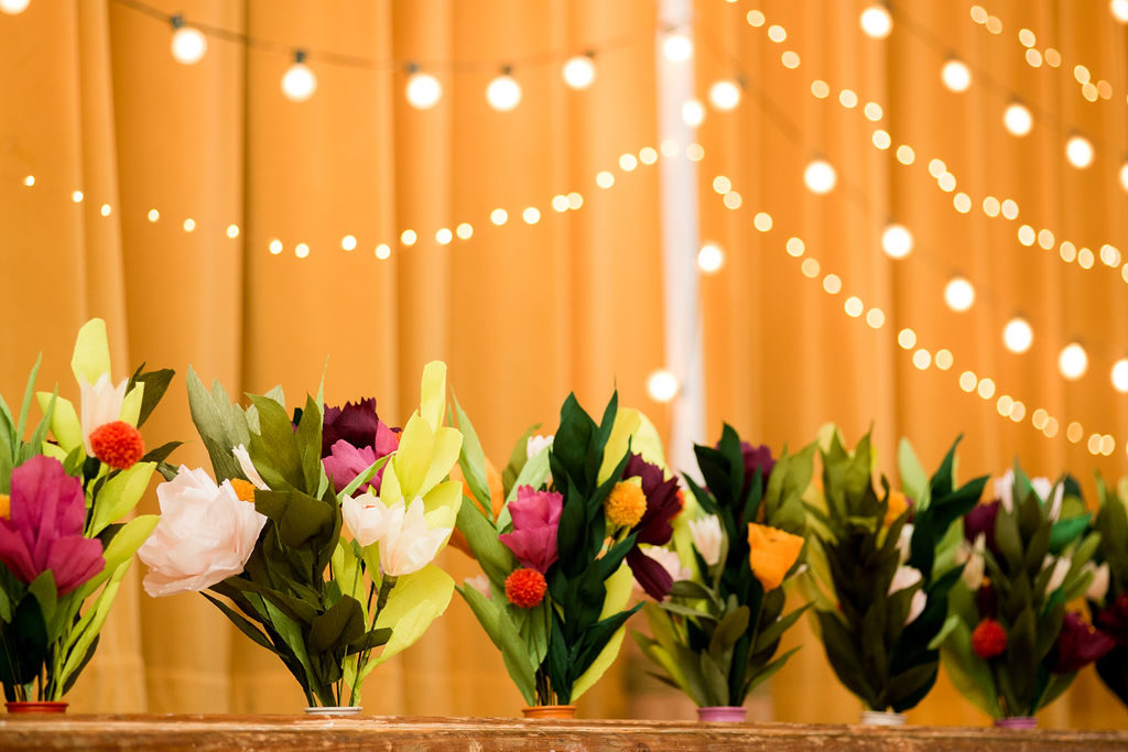 Vases of very realistic-looking paper flowers in shades of pinks and purples, with green leaves, in focus against a backdrop of gold fabric with swags of twinkly lights.