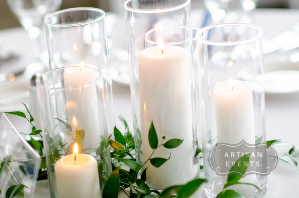 Tall white pillar candles in white glass holders surrounded by greenery.