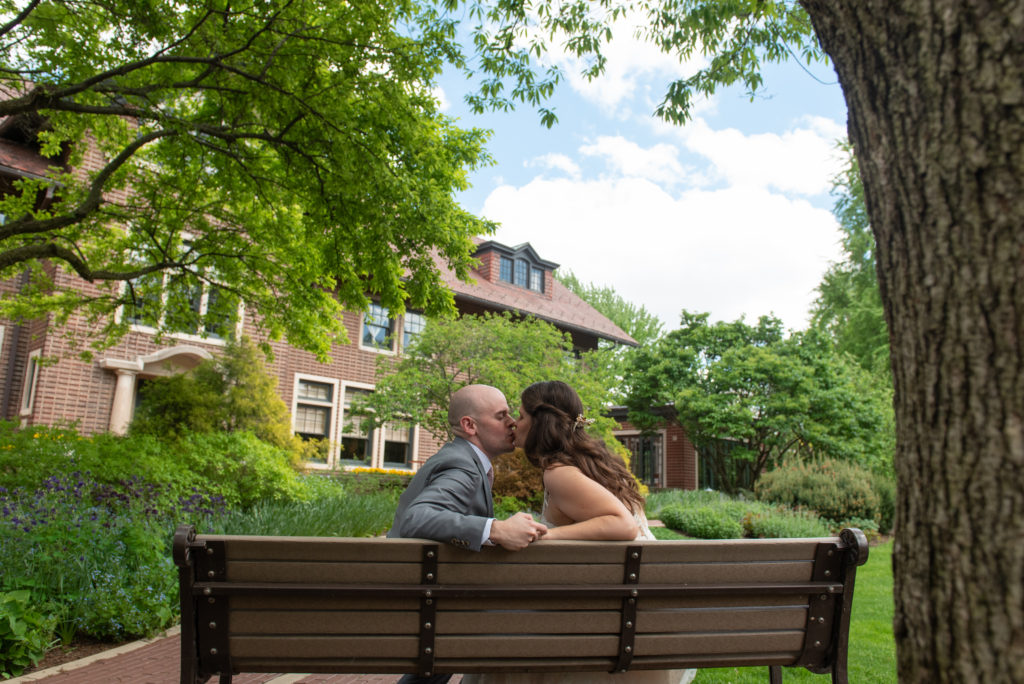 A couple kissing each other while sitting on a bench in front of a brick mansion with large grounds.