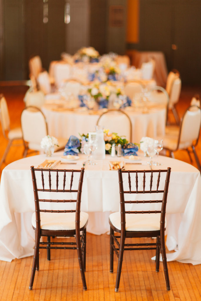A half-moon table draped in white with two wooden chairs in front of it. Flowers in shades of pink and yellow, and favors in blue, are seen on the table. In the distance are banquet tables similarly done. Everything is a little out of focus.