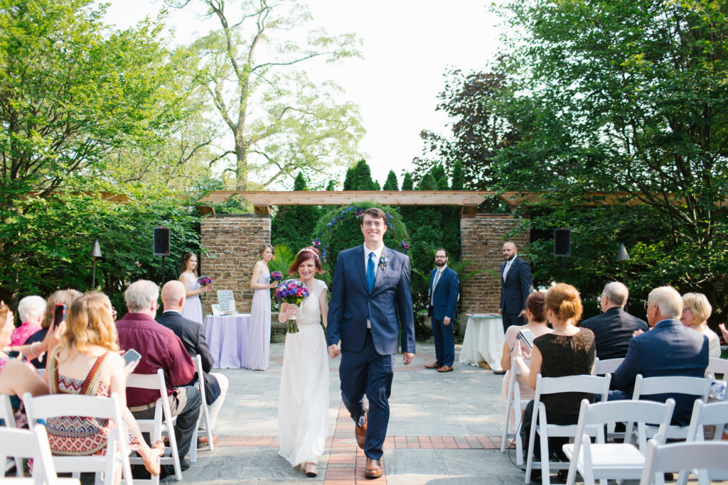 Bride and groom walk back up the aisle in an outdoor area, with a few guests seated in chairs on either side of the aisle.