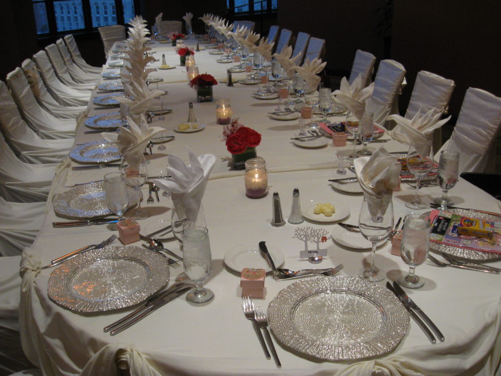 A long table, seen from the end, dressed in white linens with silver chargers at each place setting and a few flowers and candles down the length of it.